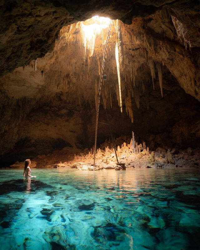 A woman resting in the turquoise water of an underground cenote cave, daylight falling through an opening onto stalactites